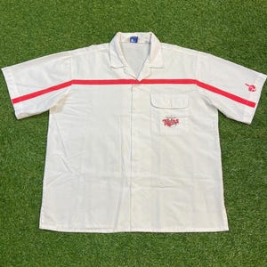 Pode incluir: Camisa branca de manga curta com listras vermelhas no peito e nas mangas. Possui um pequeno bolso com o logotipo "Twins" em vermelho. A camisa tem uma gola clássica e um ajuste relaxado.