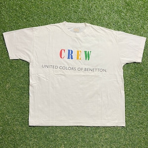 May include: A white t-shirt with the word "CREW" in red, blue, yellow, and green letters. Below the word "CREW" is the text "UNITED COLORS OF BENETTON." The shirt is laid flat on a green surface.