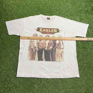 Könnte beinhalten: Weißes T-Shirt mit einer Grafik der Band Eagles und dem Text "Hell Freezes Over". Das Bild zeigt die Bandmitglieder auf einem Gruppenfoto. Das T-Shirt besteht aus einem weichen, bequemen Material.