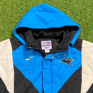 Peut inclure: Veste &agrave; capuche bleue, noire et blanche avec le logo des Panthers de la Caroline. La veste porte un logo Reebok et une &eacute;tiquette NFL Pro Line Authentic. La veste est sur une surface verte.