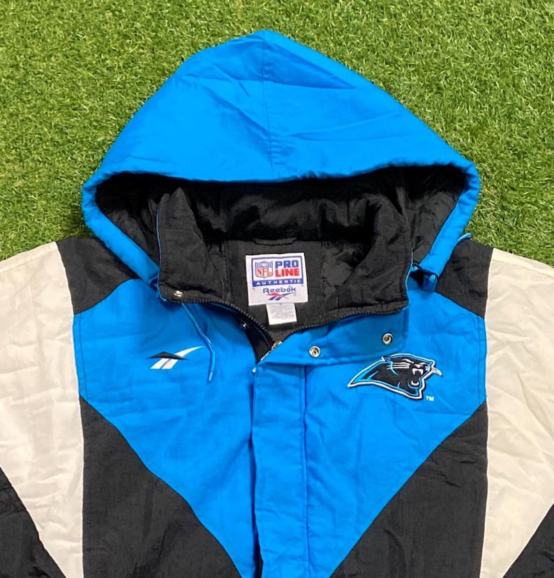 Peut inclure: Veste &agrave; capuche bleue, noire et blanche avec le logo des Panthers de la Caroline. La veste porte un logo Reebok et une &eacute;tiquette NFL Pro Line Authentic. La veste est sur une surface verte.