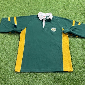 Puede incluir: Camiseta de rugby verde con rayas amarillas en las mangas y los lados. Tiene un cuello blanco y un pequeño emblema en el pecho. La camiseta de manga larga está hecha de un material suave.