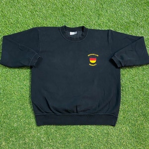 以下が含まれることがあります： 黒いスウェットシャツに、黄色と黒の刺繍でドイツ国旗と「Deutschland Germany」の文字が前面に施されています。