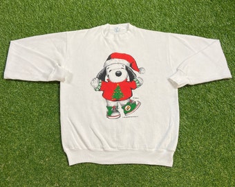 Può includere: Felpa bianca natalizia con un'immagine di Snoopy in cartone animato che indossa un cappello da Babbo Natale e tiene un albero di Natale. Il testo "Snoopy" è stampato sotto l'immagine.