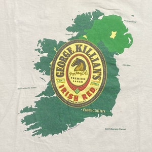 May include: A white t-shirt with a green outline of Ireland and a George Killian's Irish Red beer label. The label features a red horse and the text "George Killian's Irish Red" in a red and gold font.