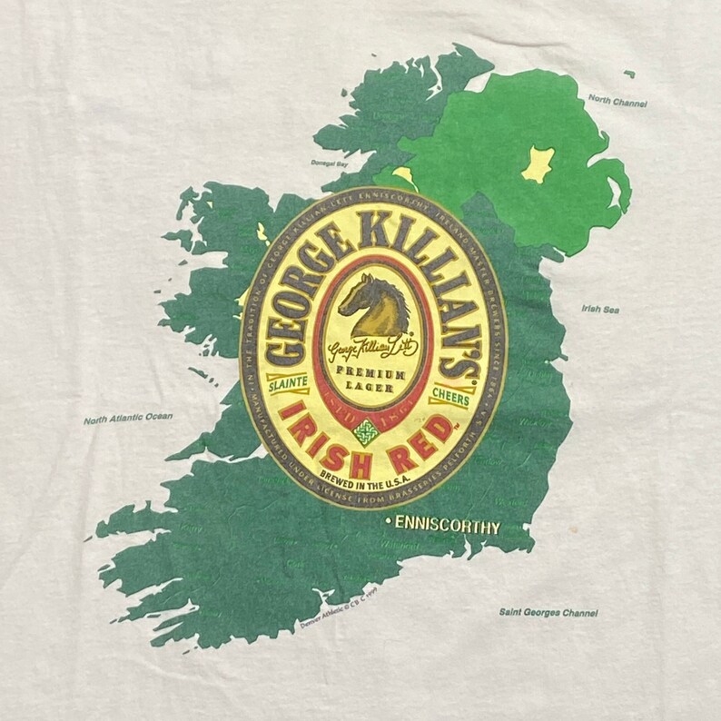 May include: A white t-shirt with a green outline of Ireland and a George Killian's Irish Red beer label. The label features a red horse and the text "George Killian's Irish Red" in a red and gold font.