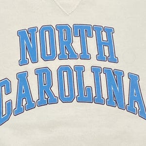 May include: Cream-colored sweatshirt with the words "NORTH CAROLINA" arched across the front in light blue block letters with a darker blue outline. The sweatshirt has a crew neck.