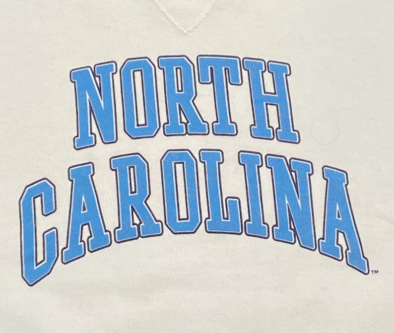 May include: Cream-colored sweatshirt with the words "NORTH CAROLINA" arched across the front in light blue block letters with a darker blue outline. The sweatshirt has a crew neck.