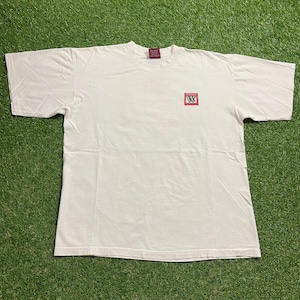 Puede incluir: Una camiseta blanca de manga corta. Un pequeño cuadrado rojo con texto blanco está impreso en la parte delantera. La camiseta está tendida sobre una superficie verde.