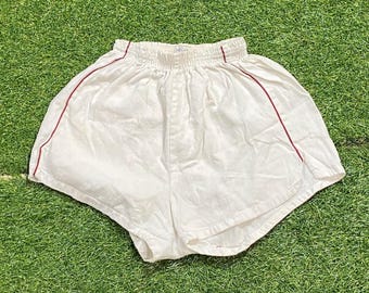 May include: White athletic shorts with a red trim along the sides and waistband. The shorts have an elastic waistband and a loose fit. The shorts are made of a lightweight fabric, perfect for sports or casual wear.