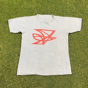 Peut inclure: T-shirt bleu clair avec un motif abstrait rouge. Le motif semble être une forme géométrique stylisée avec les lettres "NO3" incorporées. Le t-shirt est en matière souple et a un col rond.