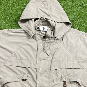 Pu&ograve; includere: Giacca con cappuccio beige Calvin Klein. La giacca presenta un cappuccio con coulisse, una chiusura con bottoni e due tasche anteriori con cerniere. Il logo Calvin Klein &egrave; visibile all'interno del cappuccio.