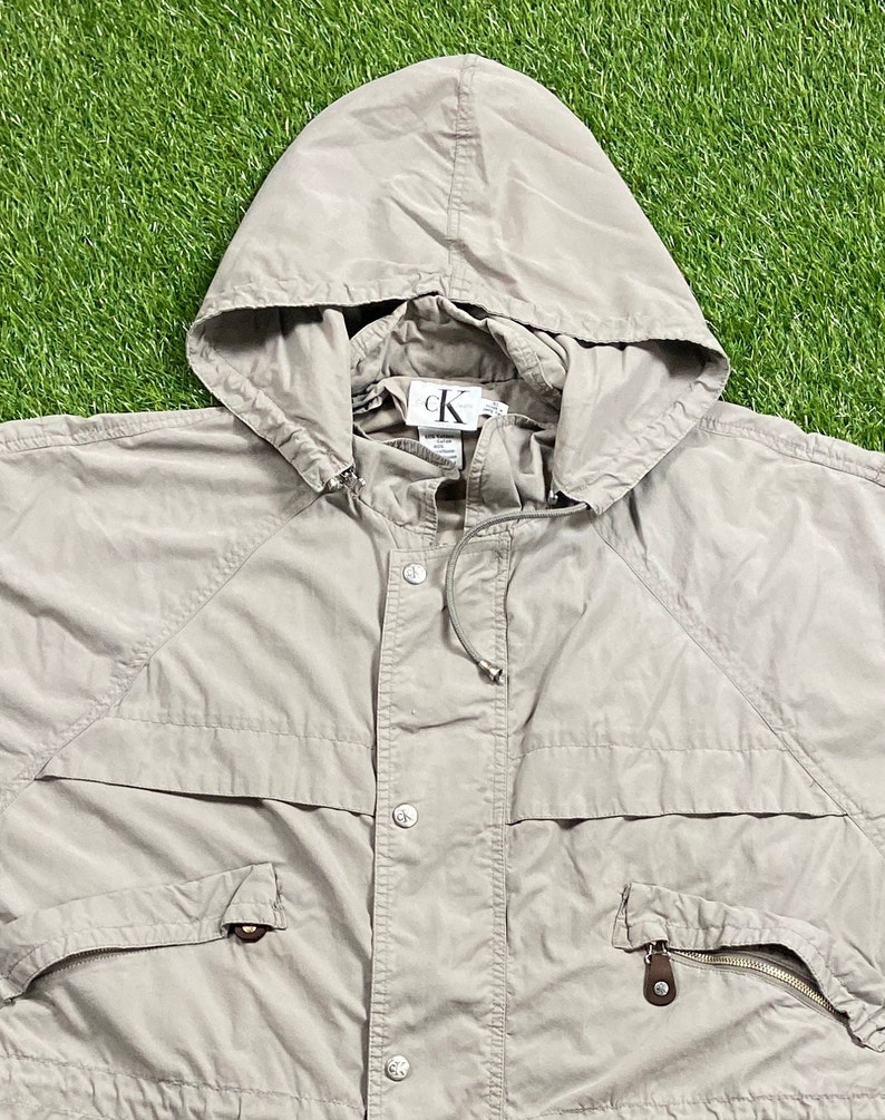 Pu&ograve; includere: Giacca con cappuccio beige Calvin Klein. La giacca presenta un cappuccio con coulisse, una chiusura con bottoni e due tasche anteriori con cerniere. Il logo Calvin Klein &egrave; visibile all'interno del cappuccio.