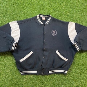 May include: A black and grey varsity jacket with a white stripe around the collar and cuffs. The jacket has a button closure and two pockets. The jacket has a logo on the left chest that says "Hollywood Stars".