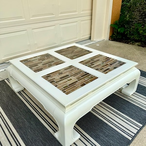 May include: A square coffee table with a white frame and four inset panels featuring a mosaic of rectangular glass tiles in shades of brown, tan, and gray. The table has curved legs and a smooth, painted finish on the wooden structure.