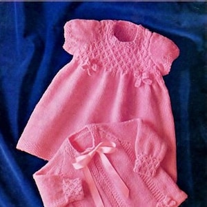 Könnte beinhalten: Ein rosa gestricktes Baby-Kleid und Cardigan-Set mit einer Schleife aus Satinband. Das Kleid hat ein strukturiertes Muster und der Cardigan ein einfaches Strickmuster.  FROM JOJO'S SECRET ATTIC.