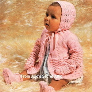Könnte beinhalten: Ein rosa gestricktes Baby-Outfit mit passendem Mützchen, Cardigan und Stiefeln. Das Outfit hat ein zartes Strickmuster und eine Schleife am Hals.