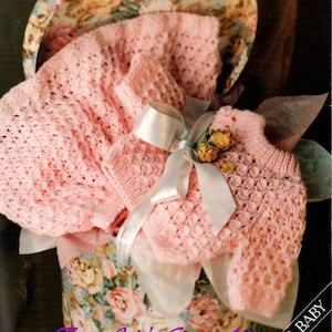 May include: A pink knitted baby sweater and hat set in a floral gift box. The set is made with a delicate stitch pattern and is tied with a white satin ribbon. The box is decorated with a floral pattern and the text "From Jojo's Secret Attic" is printed on the bottom.
