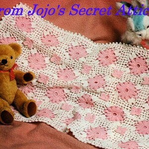 May include: A white crocheted baby blanket with pink flower accents. A brown teddy bear and a blue bunny are lying on the blanket.
