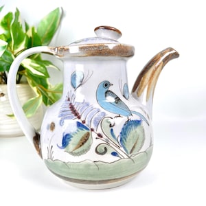 May include: White ceramic teapot with a lid, handle, and spout. The teapot is decorated with a blue bird, butterflies, and floral designs in blue, green, and brown. The base has a green border.