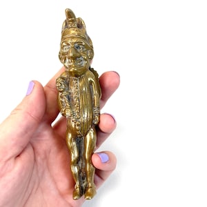 May include: A vintage brass figurine of a whimsical figure with a hat, holding a smaller figure. The detailed sculpture has a golden hue and is held in a hand against a white background. The figurine has a smiling face and is wearing a buttoned outfit.