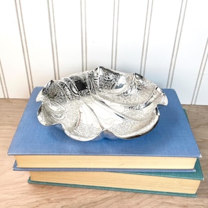 May include: A silver-colored, ornate bowl with a scalloped edge, resting on top of two blue books. The bowl features detailed floral and scrollwork patterns. The books have yellowed pages and are stacked on a wooden surface.