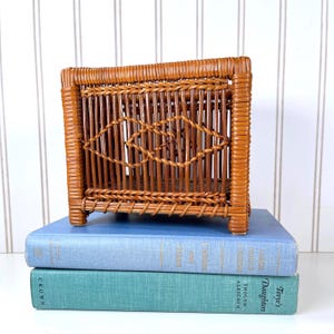 May include: A small, rectangular wicker basket with a diamond-shaped design in the center. The basket is a warm brown color and sits atop two vintage books with blue and teal covers. The background is a white wall with vertical stripes.