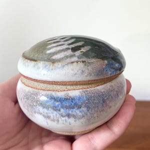 Pode incluir: Um pequeno pote de cerâmica redondo com tampa, apresentando um esmalte manchado em tons de branco, azul e verde terroso. A peça tem um acabamento liso e brilhante com bordas de argila crua visíveis onde a tampa encontra a base do recipiente.