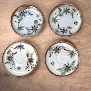 Puede incluir: Cuatro platillos de cerámica con un diseño floral. Cada platillo presenta una depresión central y está decorado con flores azules y marrones, hojas verdes y un borde marrón. Los platillos están dispuestos sobre una superficie de madera marrón claro.