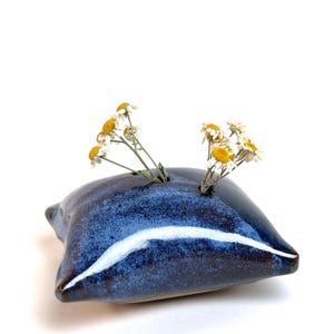 Puede incluir: Un jarrón de cerámica hecho a mano con un esmalte azul oscuro y reflejos blancos. El jarrón tiene una forma única, similar a una almohada, y contiene pequeñas flores secas amarillas y blancas. Un objeto decorativo.
