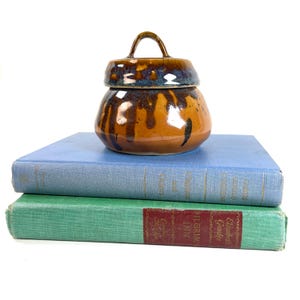 May include: A ceramic jar with a lid, featuring a brown and blue glaze, sits atop two vintage books. The jar has a handle on the lid. The books are light blue and green.
