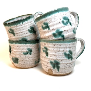 Puede incluir: Cuatro tazas de cerámica apiladas. Son blancas con un patrón moteado y diseños de hojas turquesas. Los bordes y las asas también son turquesas. Las tazas tienen un aspecto rústico y hecho a mano.