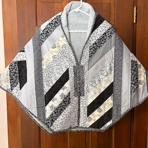 May include: A quilted cape in a patchwork design, featuring a mix of black, white, and gray fabrics. The cape has a gray collar and is hanging on a white hanger against a wooden door.