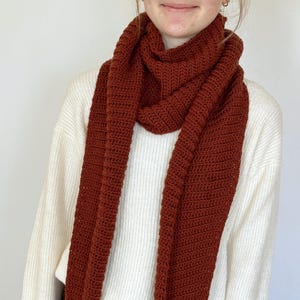 May include: A rust-colored, crocheted scarf draped over a white sweater. The scarf is made of a thick yarn, with a ribbed texture. The scarf is long and wide, suitable for wrapping around the neck. The overall style is cozy and handmade.
