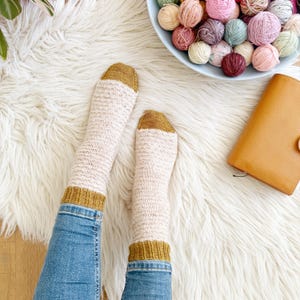 May include: A pair of white and mustard yellow knitted socks with a textured pattern. The socks are being worn by someone wearing blue jeans.