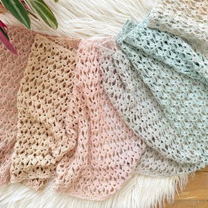 May include: Four crocheted shawls in shades of pink, beige, and gray are arranged on a white faux fur surface. The shawls have an open, lacy weave. A green plant with red stems is visible in the upper left corner. The shawls are likely for fashion or home decor.