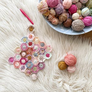 Op de afbeelding: Een verzameling garenballen in verschillende pastelkleuren, waaronder roze, paars en beige, is gerangschikt in een blauwe kom. Een roze haaknaald ligt naast een gehaakt zeshoekig stuk. Drie kleine garenballen zijn ook zichtbaar.