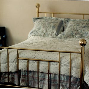 May include: A brass bed frame with spherical finials on the headboard and footboard posts. The bed is dressed in a white comforter featuring a small blue floral pattern, a matching bed skirt, and two pillows with a larger blue floral print.