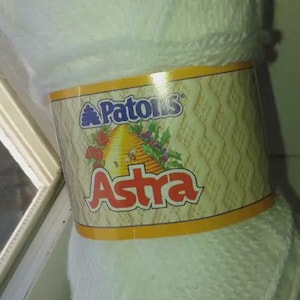 May include: A skein of white yarn wrapped in a paper label featuring the Patons brand logo, a beehive illustration, and the text Astra in large red letters. The label has a textured, light-coloured background with orange borders at the top and bottom.