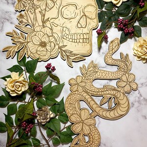 以下が含まれることがあります： 木製のスカルとヘビの切り抜き、花の模様。スカルは花と葉で飾られています。ヘビは巻き付いており、花で飾られています。人工の花と緑が作品を囲んでいます。