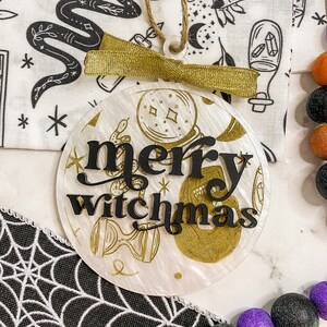 May include: A white round ornament with a gold glitter finish and black lettering that reads "merry witchmas". The ornament is decorated with gold glitter and black illustrations of a snake, a hand holding a crystal, and a crescent moon.