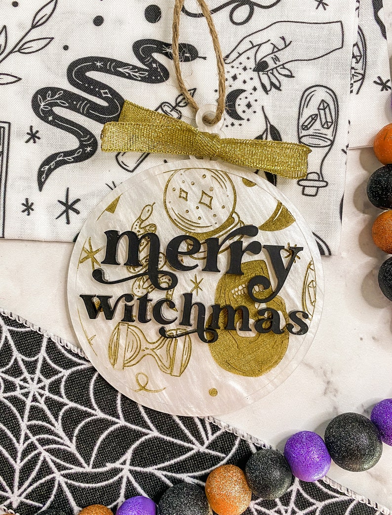 May include: A white round ornament with a gold glitter finish and black lettering that reads "merry witchmas". The ornament is decorated with gold glitter and black illustrations of a snake, a hand holding a crystal, and a crescent moon.