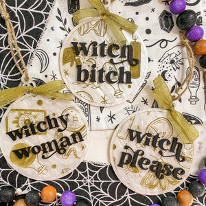 May include: Three round, white, decorative ornaments with gold accents and black lettering. The ornaments read "Witchy Woman", "Witch Bitch", and "Witch Please".