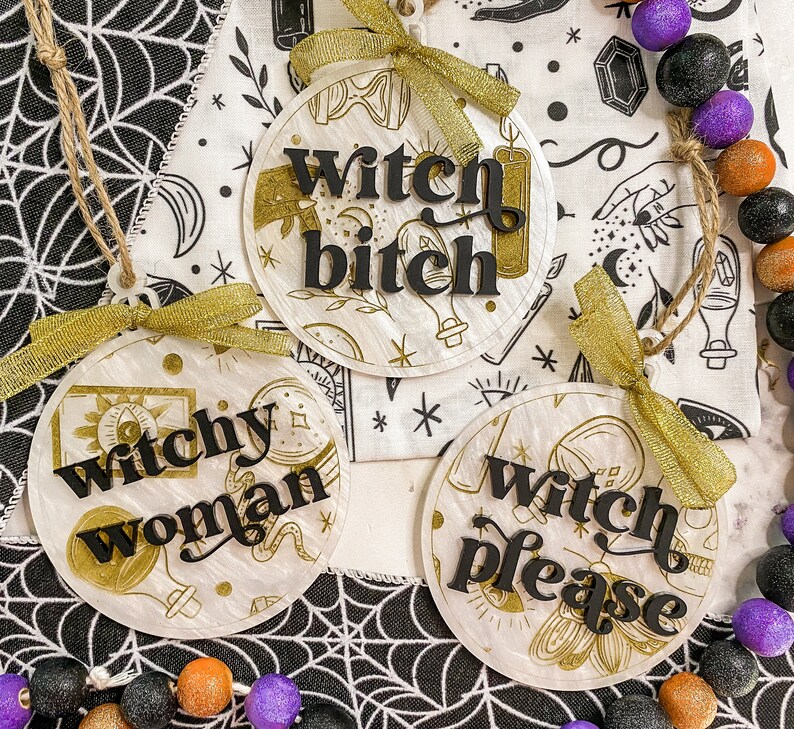 May include: Three round, white, decorative ornaments with gold accents and black lettering. The ornaments read "Witchy Woman", "Witch Bitch", and "Witch Please".