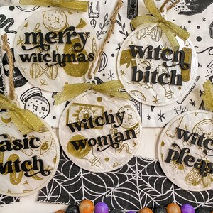 May include: Five round, white, decorative ornaments with gold accents and black lettering. The ornaments feature the phrases "merry witchmas", "witchy woman", "basic witch", "witch bitch", and "witch please".