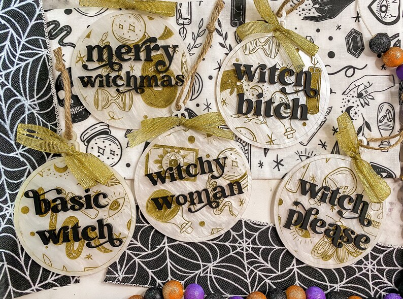 May include: Five round, white, decorative ornaments with gold accents and black lettering. The ornaments feature the phrases "merry witchmas", "witchy woman", "basic witch", "witch bitch", and "witch please".