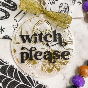 May include: A white round ornament with gold glitter and a black inscription that reads "witch please". The ornament is tied with a gold ribbon and has a string for hanging.