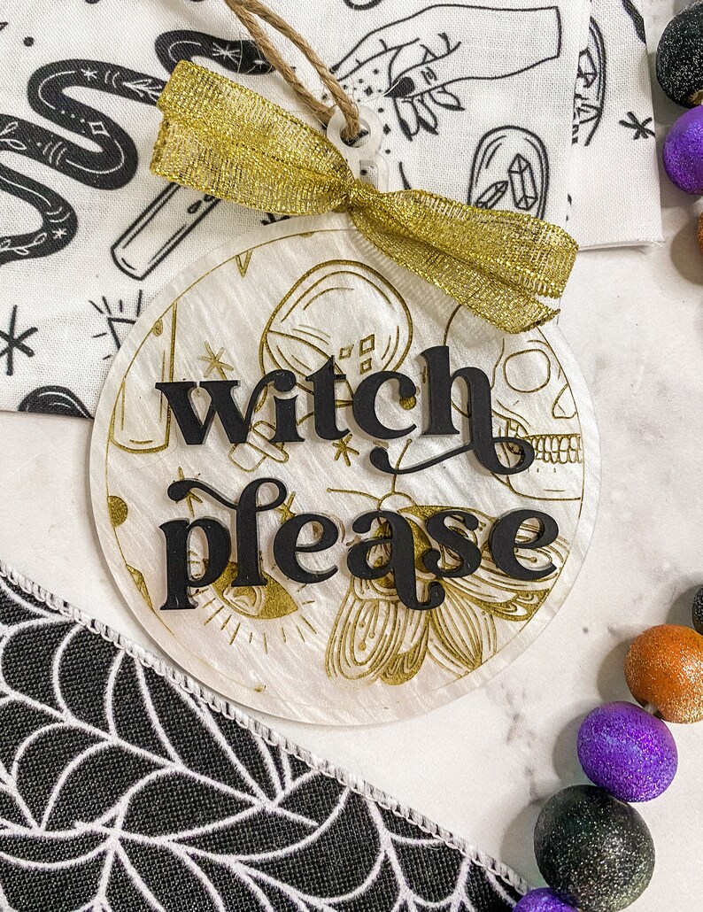 May include: A white round ornament with gold glitter and a black inscription that reads "witch please". The ornament is tied with a gold ribbon and has a string for hanging.