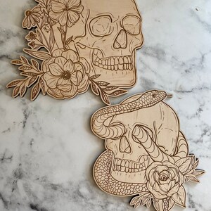 以下が含まれることがあります： 2つの木製のスカルカットアウト。1つのスカルは、花や葉を含む花の要素で飾られています。もう1つのスカルには、バラと葉が付いたヘビが巻き付いています。カットアウトは薄茶色です。