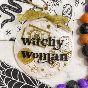 May include: A white round ornament with a gold glitter finish and black lettering that reads "Witchy Woman". The ornament has a gold ribbon and a twine hanger.